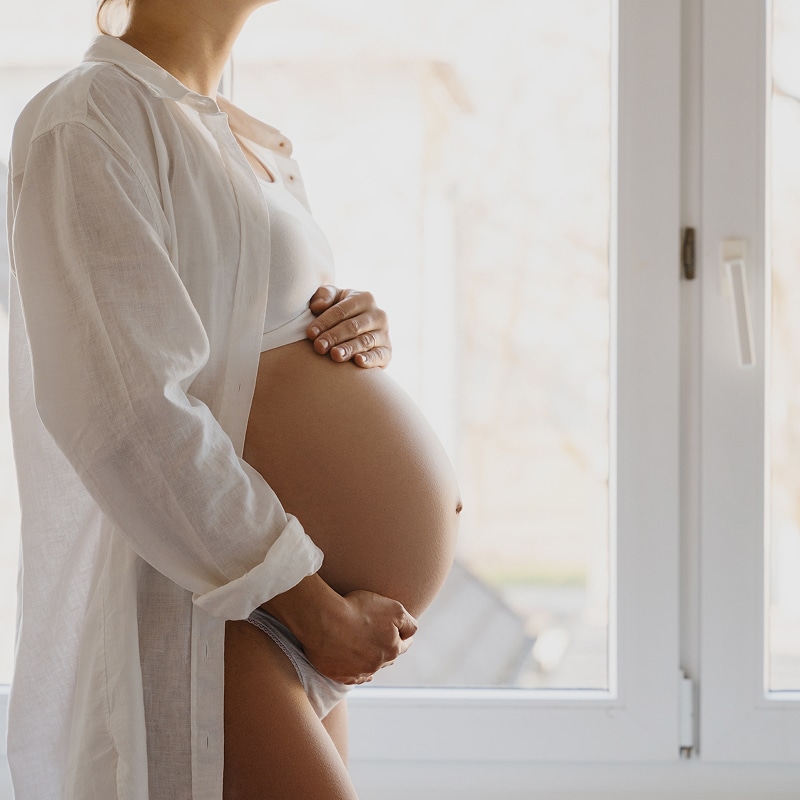 Femme enceinte tenant son ventre devant une fenêtre Femme enceinte en chemise blanche tenant son ventre arrondi devant une fenêtre lumineuse, atmosphère sereine.