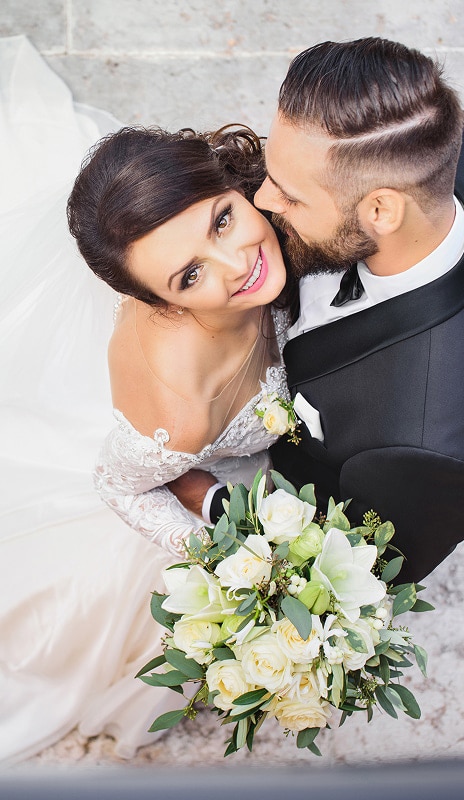 Mariage : Couple Radieux avec Bouquet de Roses Blanches Mariée souriante avec un bouquet de roses blanches, enlacée par son mari en costume, vue de dessus lors du mariage.