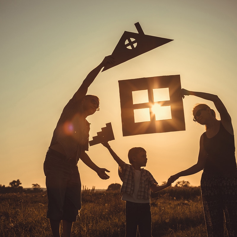 Famille et projet de maison : construire son bonheur Silhouette d'une famille heureuse formant une maison en carton au coucher du soleil, symbolisant le projet de vie.
