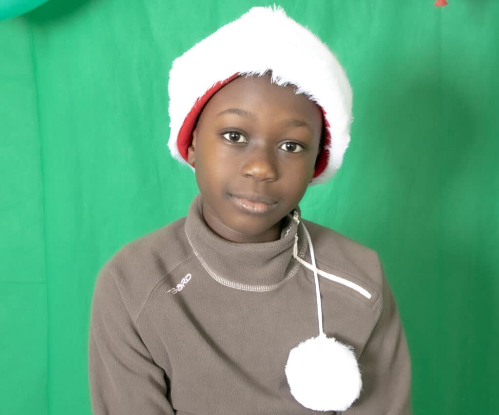 Enfant en Bonnet de Père Noël : Portrait Festif sur Fond Vert Enfant noir portant un bonnet de Père Noël et un pull Tribord marron sur fond vert pour les fêtes.