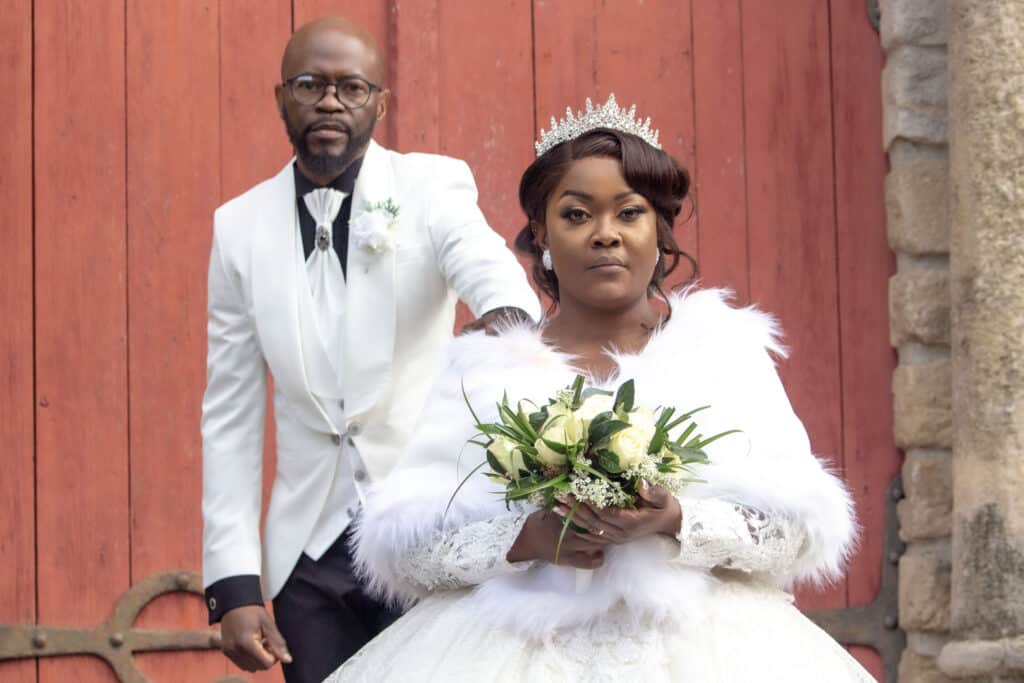 Mariage Élégant : Mariée en Blanc et Marié en Smoking Mariée couronnée avec étole en fourrure et bouquet devant un marié en smoking blanc et porte en bois rouge.