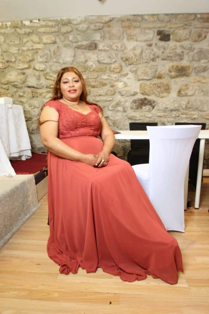 Robe de Soirée Terre Cuite : Élégance et Style Rustique Femme en robe longue terracotta avec corsage en dentelle, assise devant un mur en pierre naturelle.